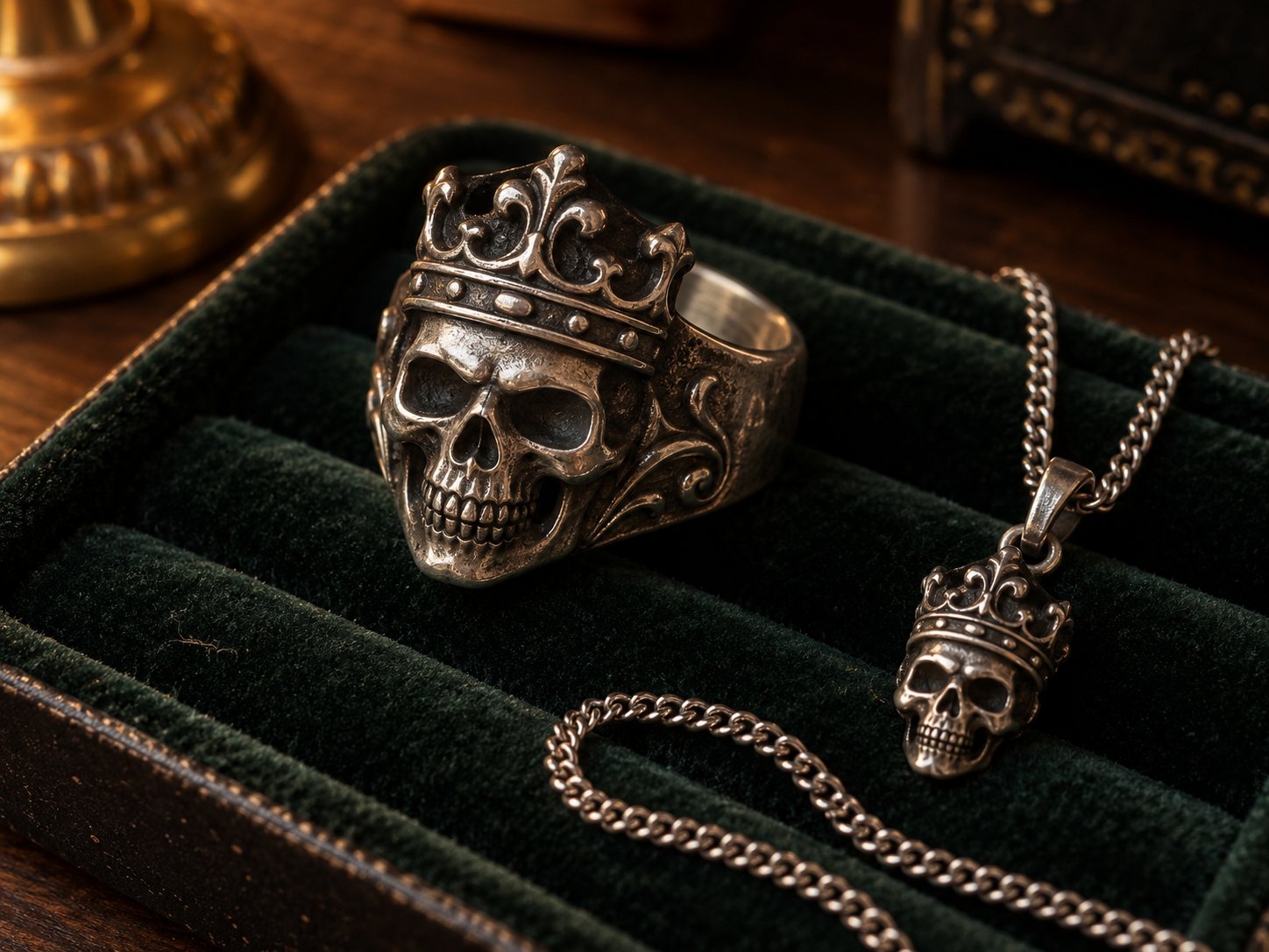 Illustration: Heavy silver ring with a crowned skull motif and a silver pendant with a crowned skull charm on a dark velvet jewelry tray