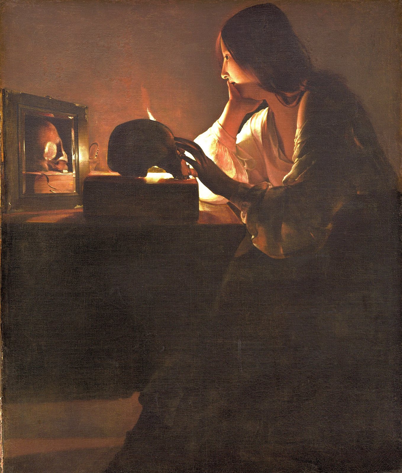 A young woman rests her chin on one hand and touches a skull lit by a single candle, with the candle and skull reflected in a mirror