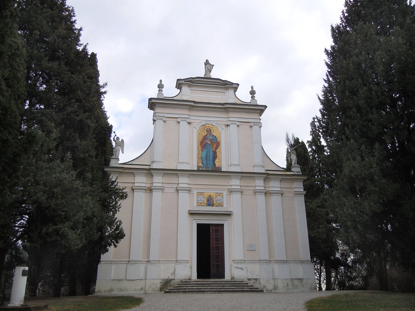 The pale pink façade of the church and ossuary chapel of San Pietro in Vincoli at Solferino, with a mosaic of a saint above the door, framed by tall dark cypress trees