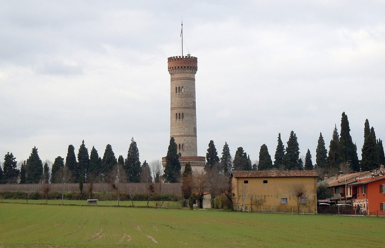 The tall, round brick tower of San Martino della Battaglia rising behind a line of cypress trees, across a green field under a grey sky