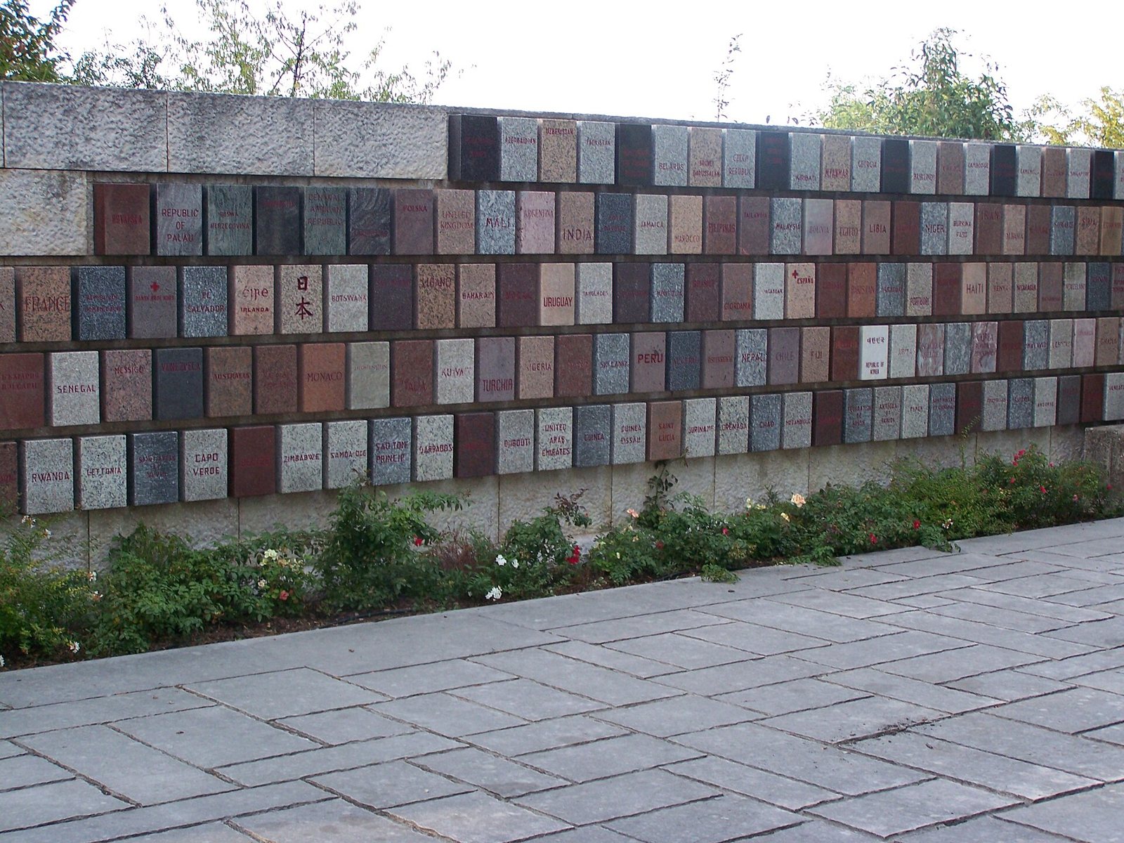 A low stone wall of small plaques in many colours and stones, each engraved with the name of a country, beside a paved walkway and a bed of flowers
