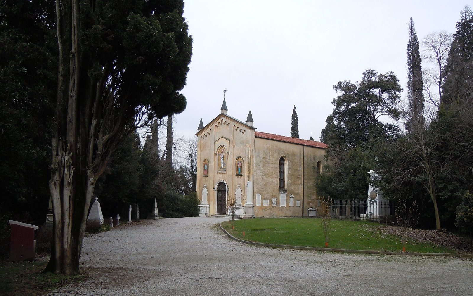A small ochre chapel with a red tiled roof and pointed turrets at the end of a gravel path among cypress trees and low grave markers at San Martino della Battaglia