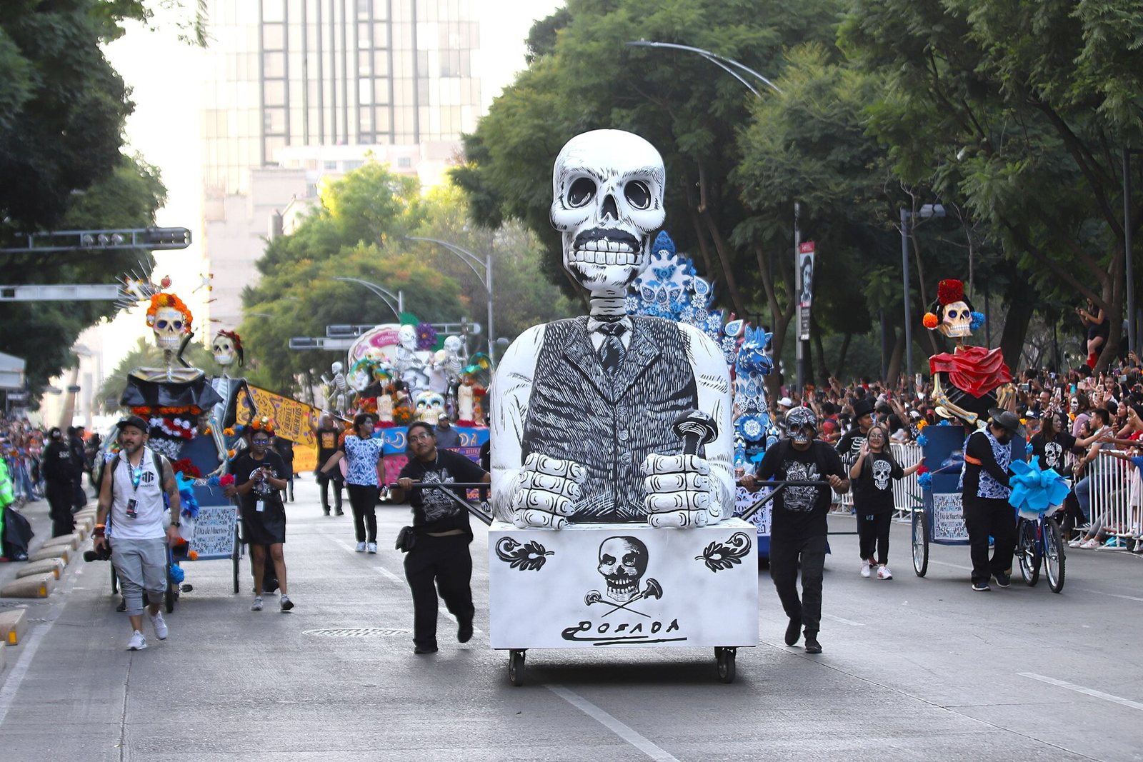 A tall white-and-black float of a skeleton in a waistcoat with clenched fists, above a skull and crossbones, pulled down a tree-lined avenue past crowds and other skeleton floats