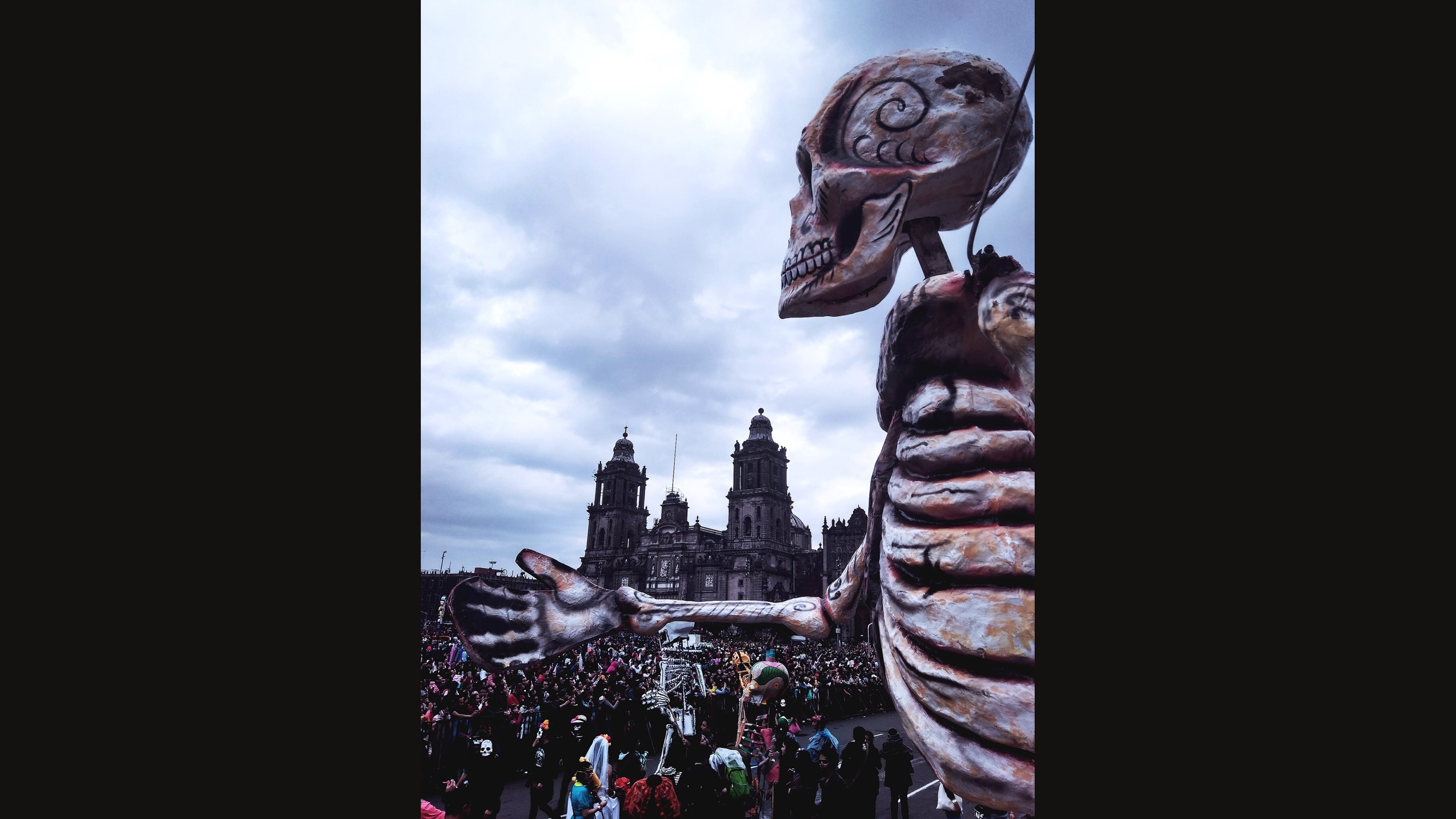 A giant papier-mache skeleton float with a swirl-patterned skull reaches an arm toward a crowd in front of the Metropolitan Cathedral under a cloudy sky