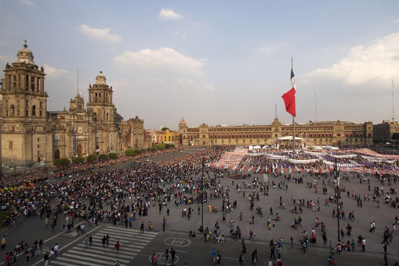 An aerial view of the Zócalo packed with crowds beside the Metropolitan Cathedral, with a large Mexican flag and the National Palace at the far side