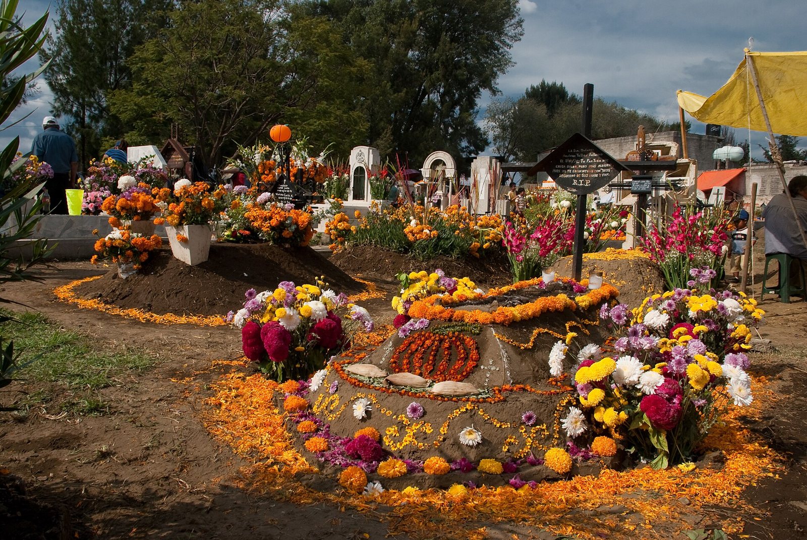 Graves in a cemetery covered in orange marigold petals, red and white flowers and candles, with a dark wooden cross and visitors among the headstones