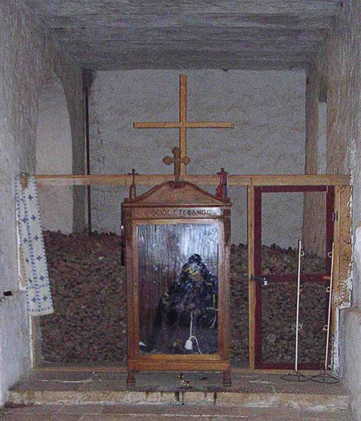 A wooden glass-fronted case holding a seated, robed figure, set in an arched niche in front of a heaped bank of bones, with a wooden cross above it