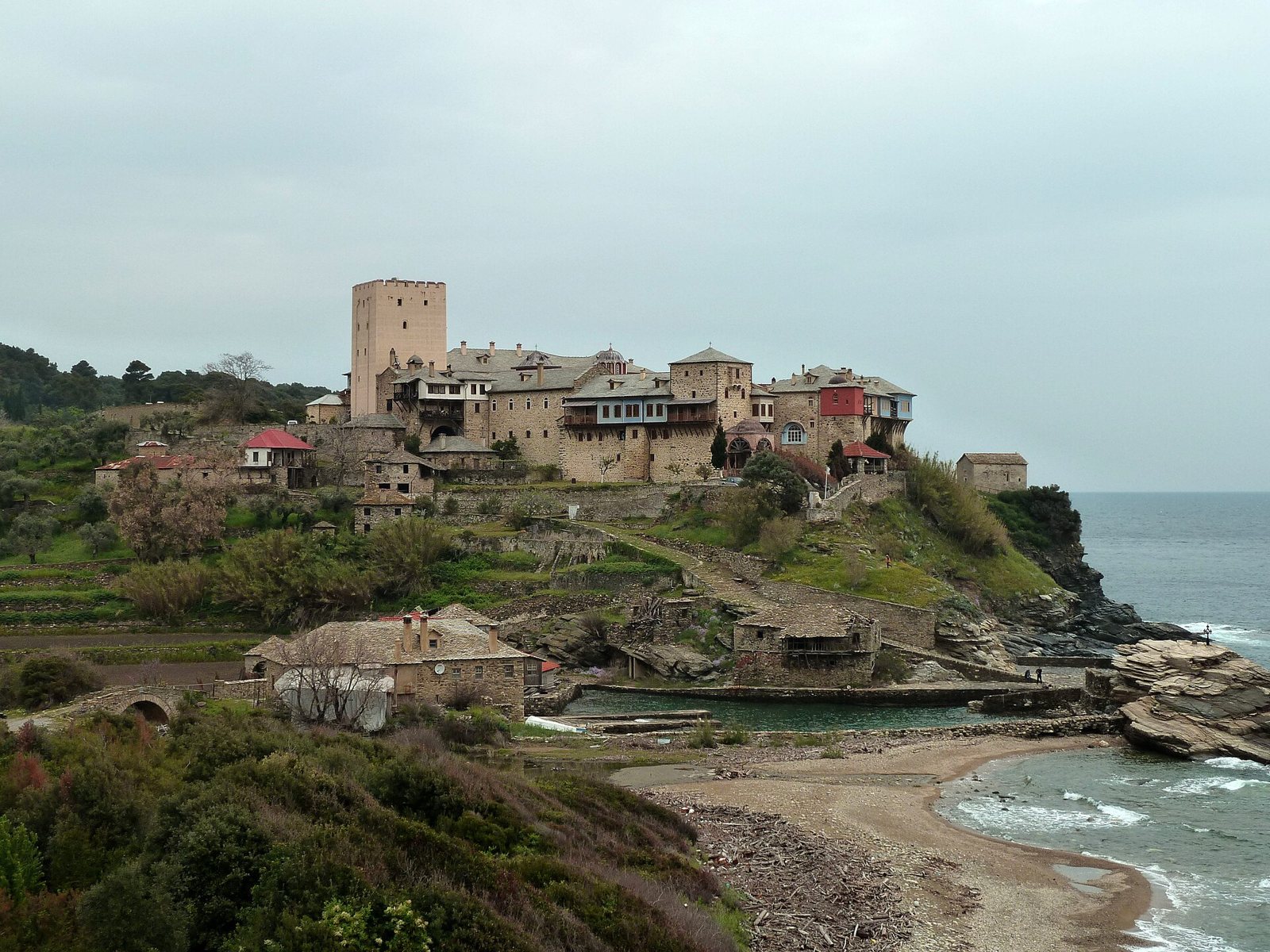 The Pantokratoros monastery on Mount Athos, a cluster of stone buildings with a tall square tower on a green headland above a small rocky cove and the sea