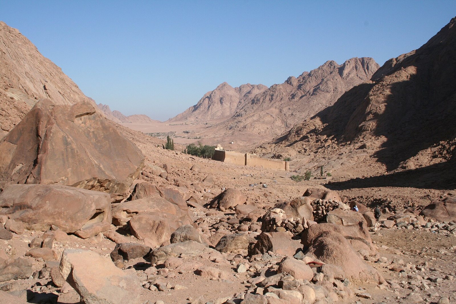 A rocky valley of boulders and red granite slopes in Sinai, with the long wall of Saint Catherine's Monastery small in the middle distance and desert mountains beyond