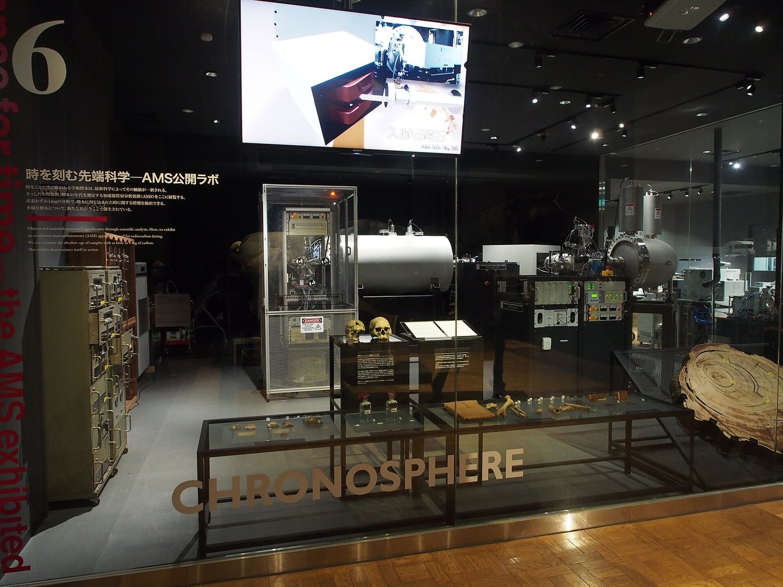 A dim museum gallery with a glass-walled radiocarbon laboratory display: a large accelerator mass spectrometer, racks of equipment, a screen above and the word CHRONOSPHERE on the glass