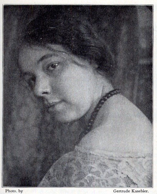 Black and white portrait photograph of Pamela Colman Smith seated and looking back over her shoulder, with dark hair, a beaded necklace and a lace collar