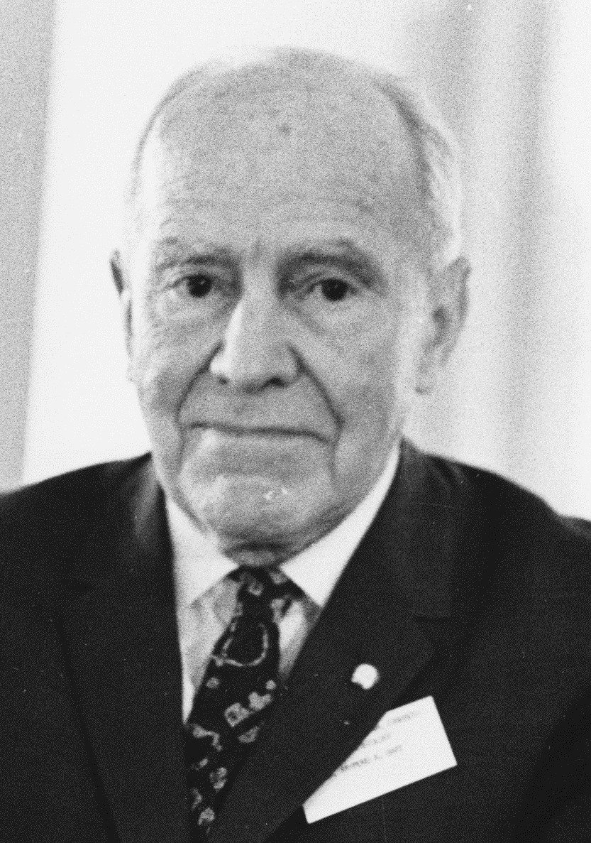 Black and white portrait of an elderly man with thin white hair, wearing a dark suit, a patterned tie and a conference name badge