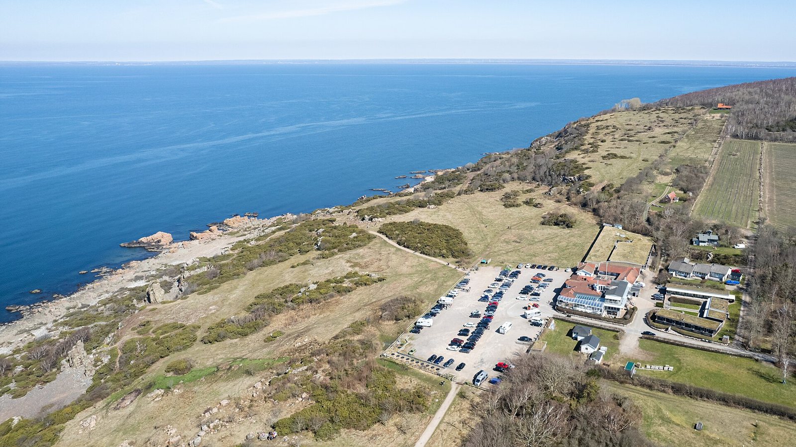An aerial view of the Hovs hallar headland in Skåne, with a rocky grass-covered coastline, a calm blue sea and a car park and buildings beside the cliff
