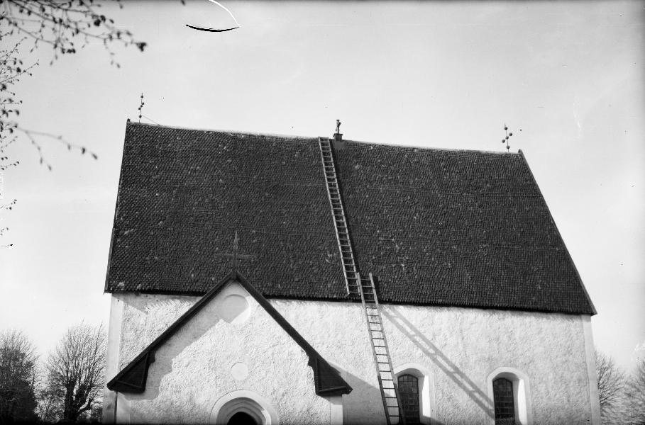A black-and-white photograph of a whitewashed stone church with a steep dark tiled roof, a ladder leaning on the roof and arched windows along the wall