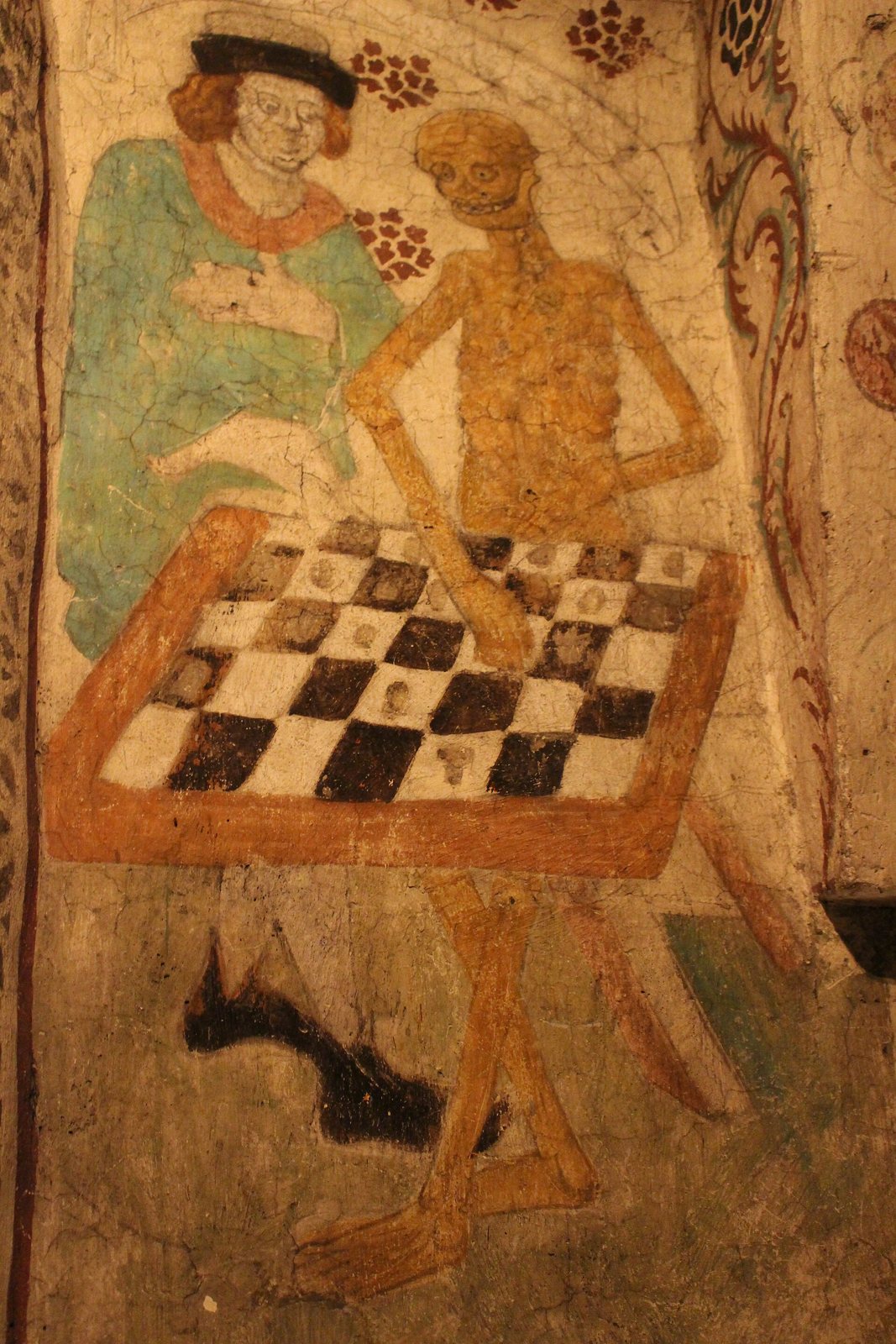 Close view of the Täby wall painting: a man in a blue-green cloak and Death, a yellow skeleton, face each other over a chessboard with pieces still visible on the squares