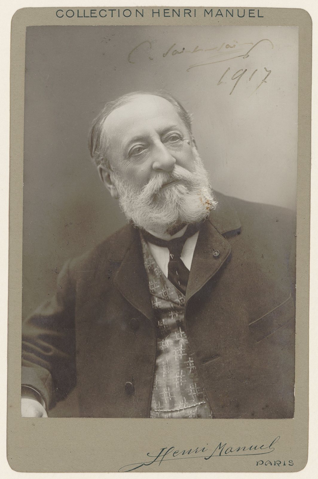 A sepia studio photograph of Camille Saint-Saëns as an elderly man with a full white beard, seated in a dark suit