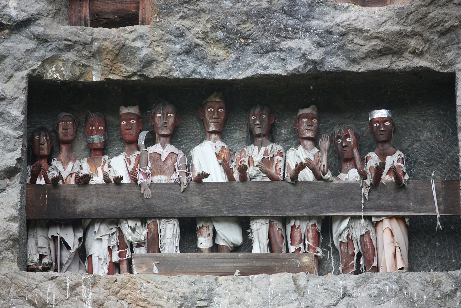 A row of carved wooden tau tau figures dressed in white cloth, standing behind a wooden rail in a balcony cut into grey rock, their painted eyes looking outward and their hands raised