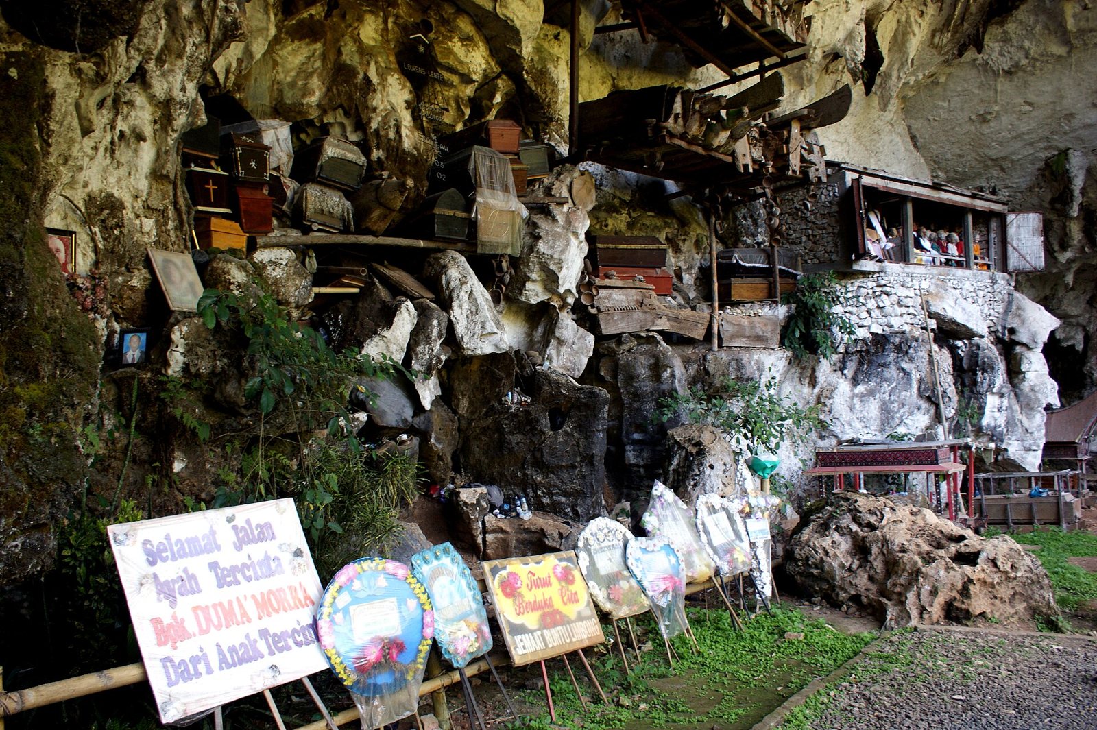 Wooden coffins and small grave houses resting on ledges along the foot of a limestone cliff at Londa, with rows of family funeral banners and flower boards standing in the grass in front