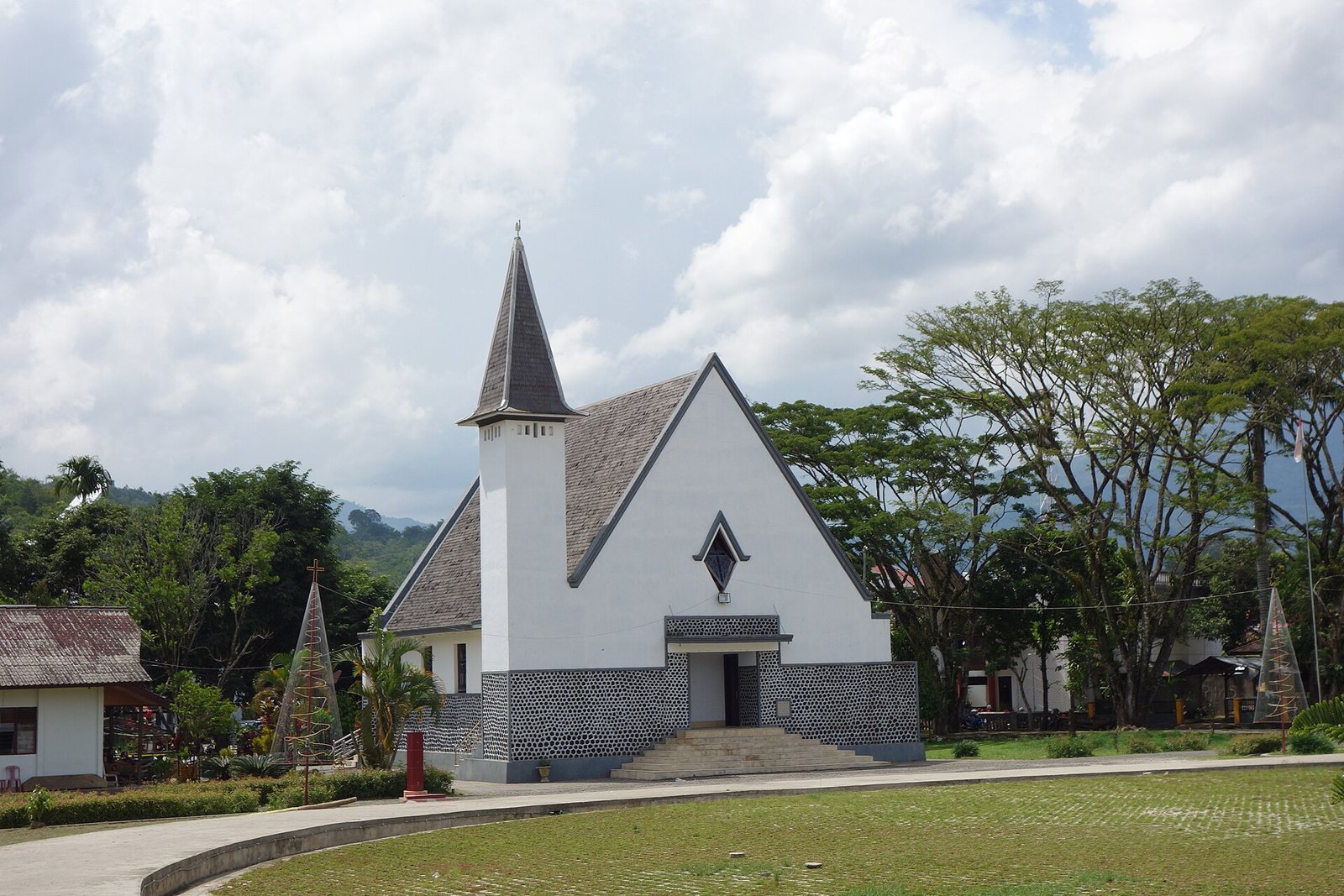 A white church with a steep grey roof and a pointed spire on a tower beside the entrance, set on a lawn with trees and green hills behind it