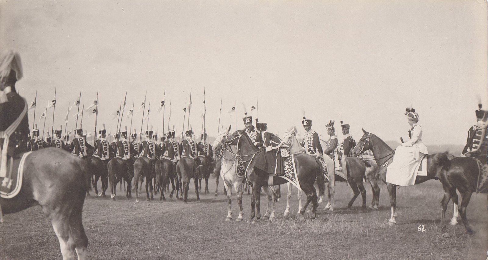 Old photograph of a parade field: mounted hussars in braided jackets and tall fur caps ride in a long line, their lance pennants marked with small skulls, while a group of riders and a woman in a pale riding dress watch in the foreground