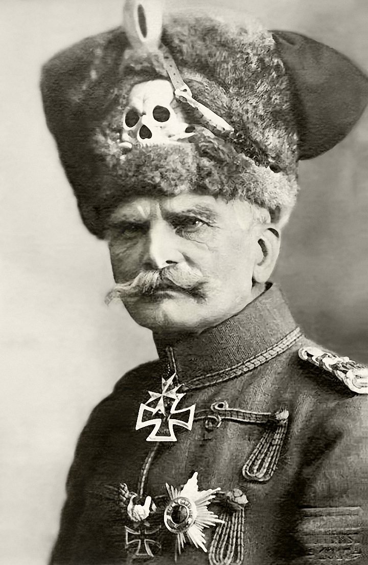 Black and white portrait of August von Mackensen, an older man with a white moustache, wearing a tall fur hussar cap with a silver skull badge and a dark braided tunic with an Iron Cross at the collar