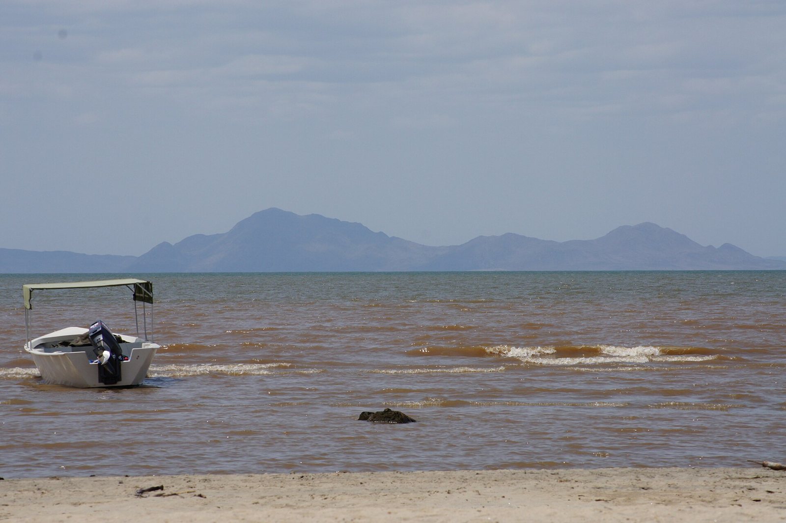 Lake Turkana in northern Kenya: brown water with small waves, a boat pulled up on the sandy shore and a line of low mountains across the horizon