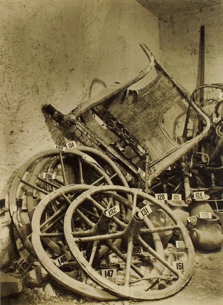 Sepia photograph of a jumble of chariot wheels, the broken frame of a chair and clay jars piled against a plastered wall, each marked with a small white numbered card