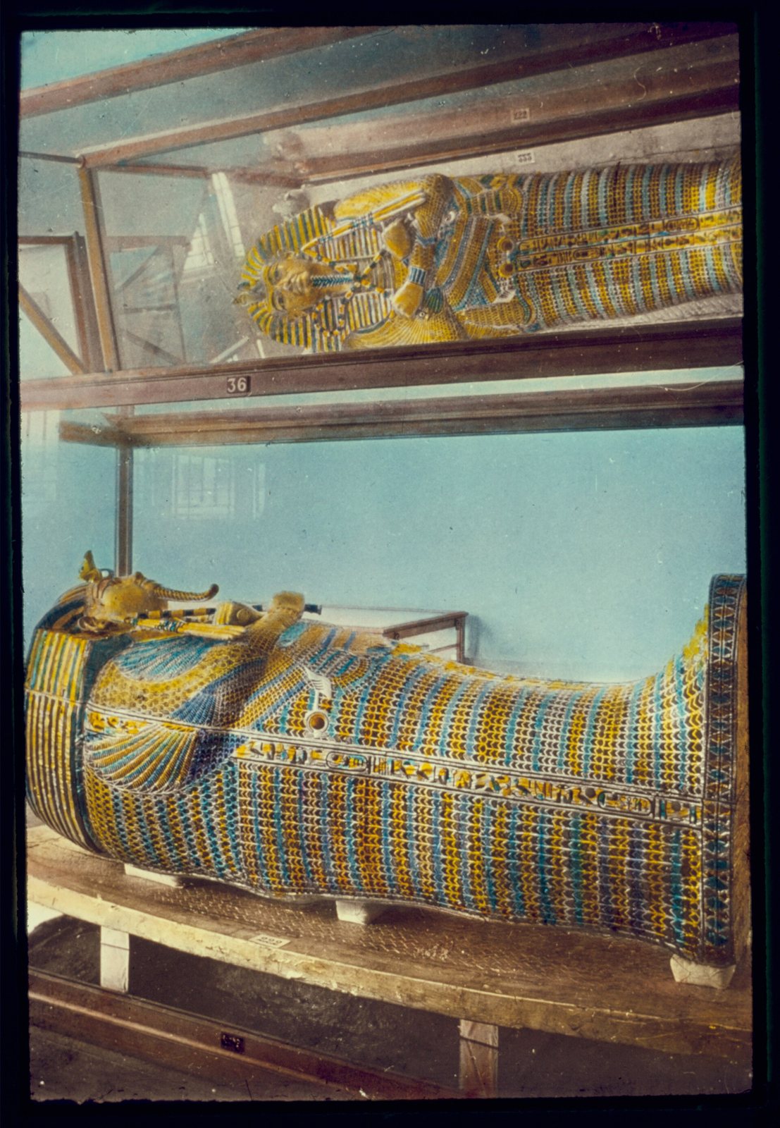 Hand-coloured slide of two gilded coffins in glass cases, covered in blue, yellow and gold feather patterns and hieroglyphs, each with a pharaoh's head, one case above the other