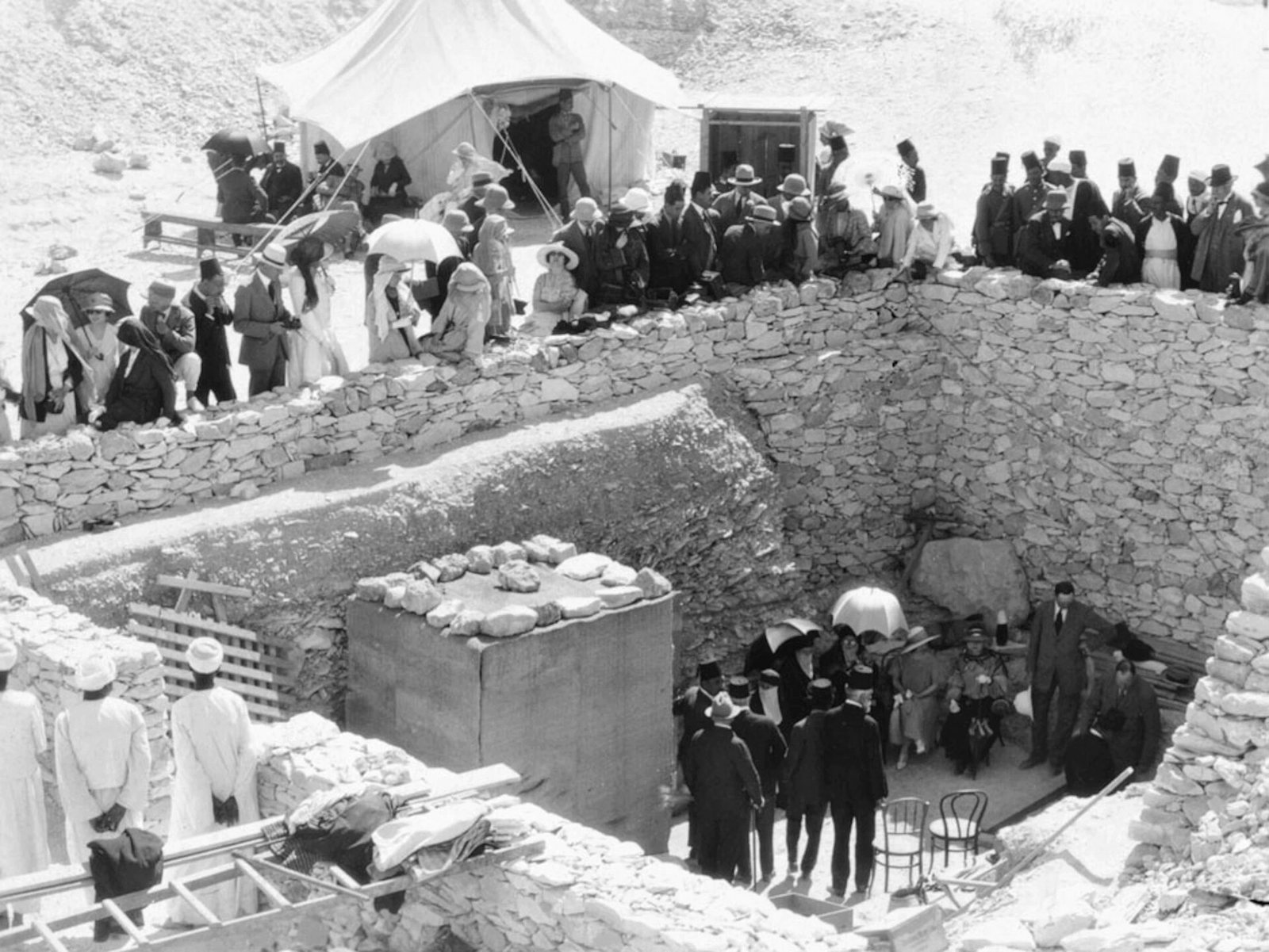 Black-and-white photograph from above of a crowd in hats, veils and fezzes standing along a dry stone wall, with a white tent above and a few dressed-up visitors sitting in the pit at the tomb entrance below