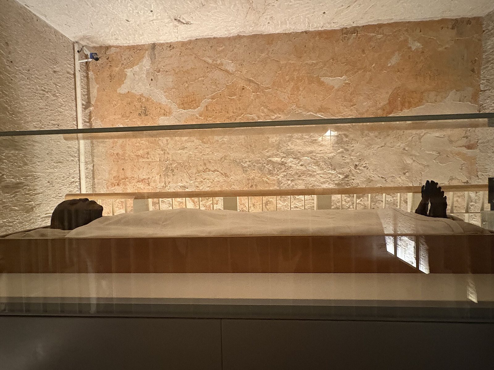Inside the tomb chamber, a mummy lying in a glass-topped wooden case: a dark head at the left end, a white linen sheet over the body and dark feet at the right end, with a pale painted rock wall behind