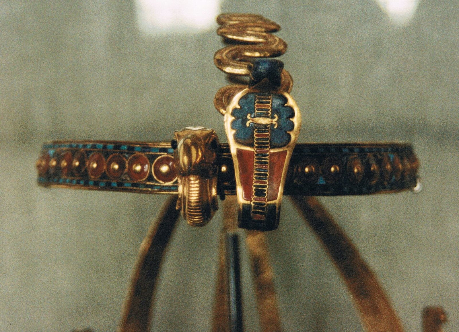 Close view of a gold diadem behind glass: a narrow band inlaid with red carnelian discs and blue and turquoise bands, with a blue-and-red inlaid cobra and a gold bird head at the front and long gold ribbon coils rising behind