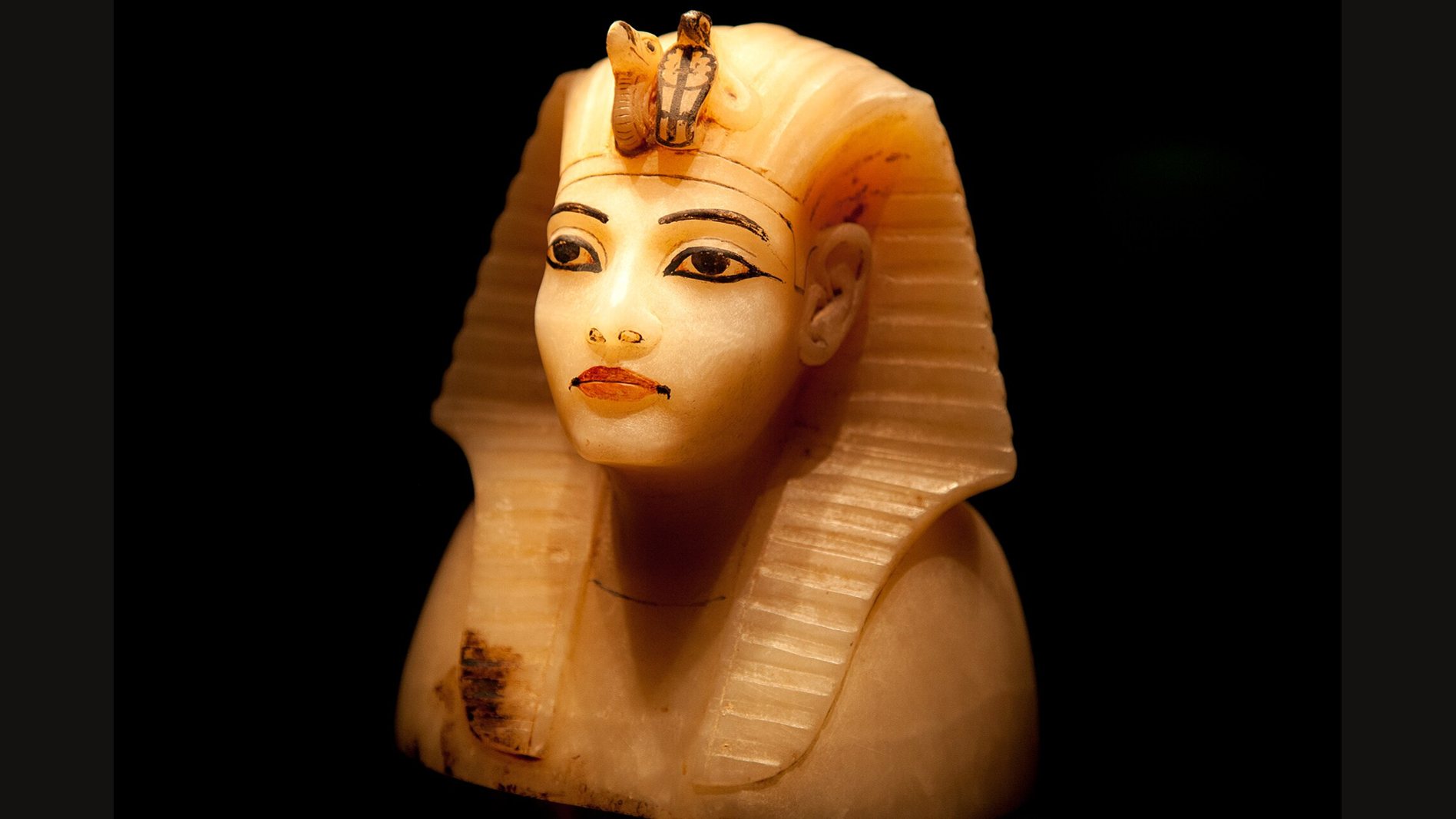 A canopic jar lid of pale alabaster carved as the head of Tutankhamun, with painted eyes and lips, a striped royal headcloth and a cobra and vulture at the brow, lit against a black background