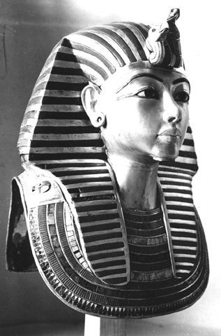 Black-and-white photograph of Tutankhamun's gold funeral mask seen from three-quarter view, showing the striped royal headcloth, a cobra and vulture at the brow, large outlined eyes and a broad beaded collar, with no beard attached