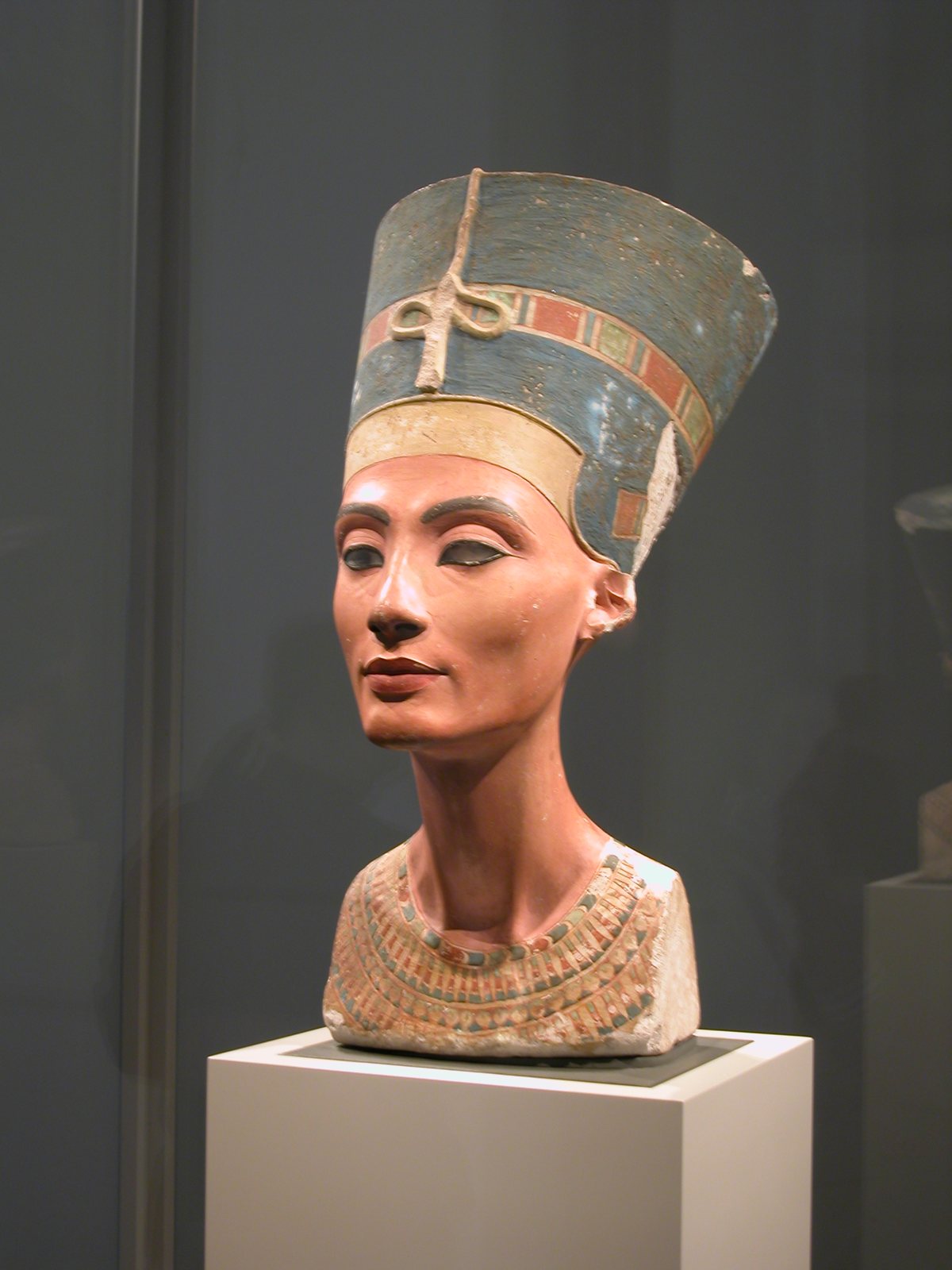 The painted limestone bust of Queen Nefertiti in a museum case: a tall flat-topped blue crown with a coloured band, a long slender neck and a broad collar of coloured beads, against a dark grey wall