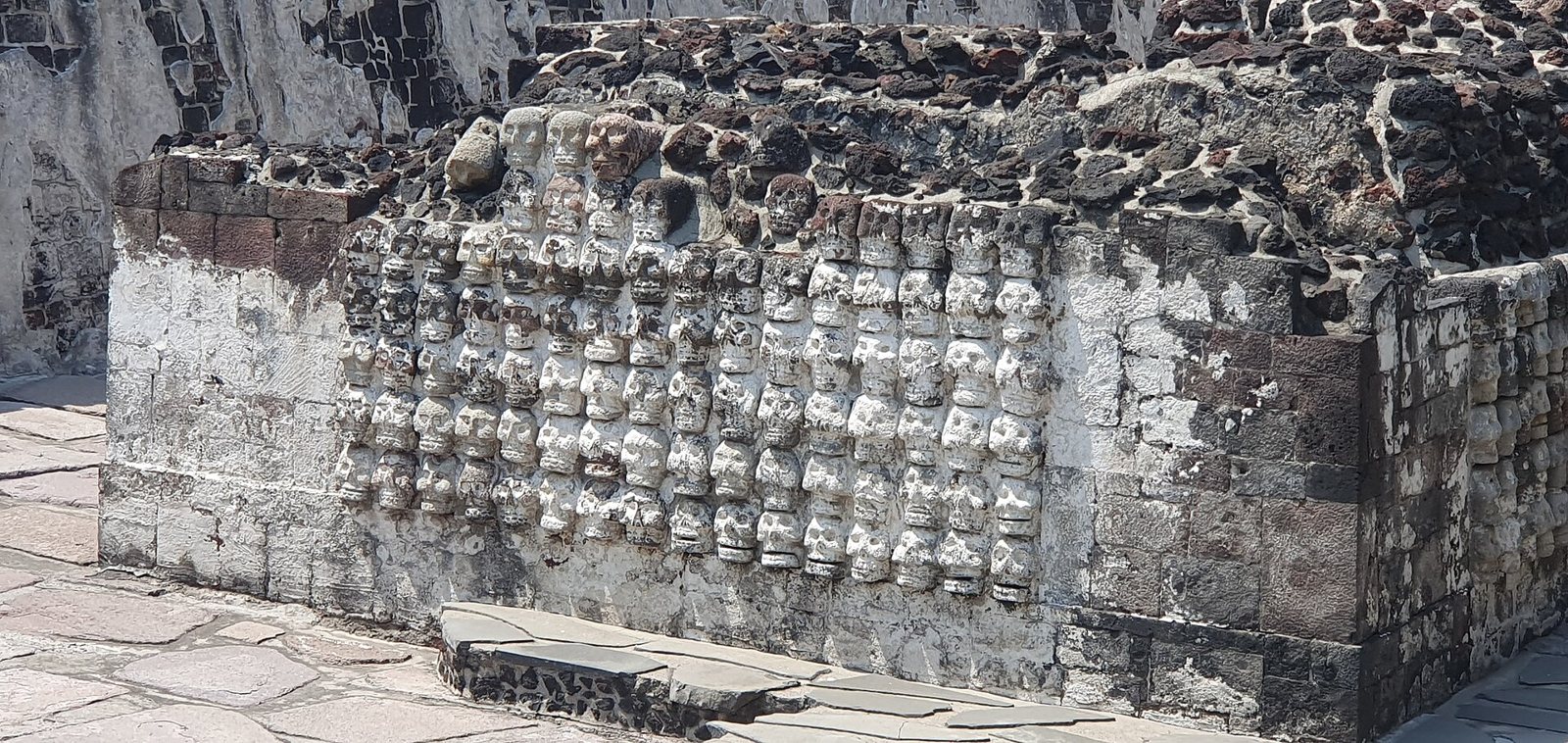 A wall of carved stone skulls in rows on a low platform at the Templo Mayor site in Mexico City, with dark volcanic stone above