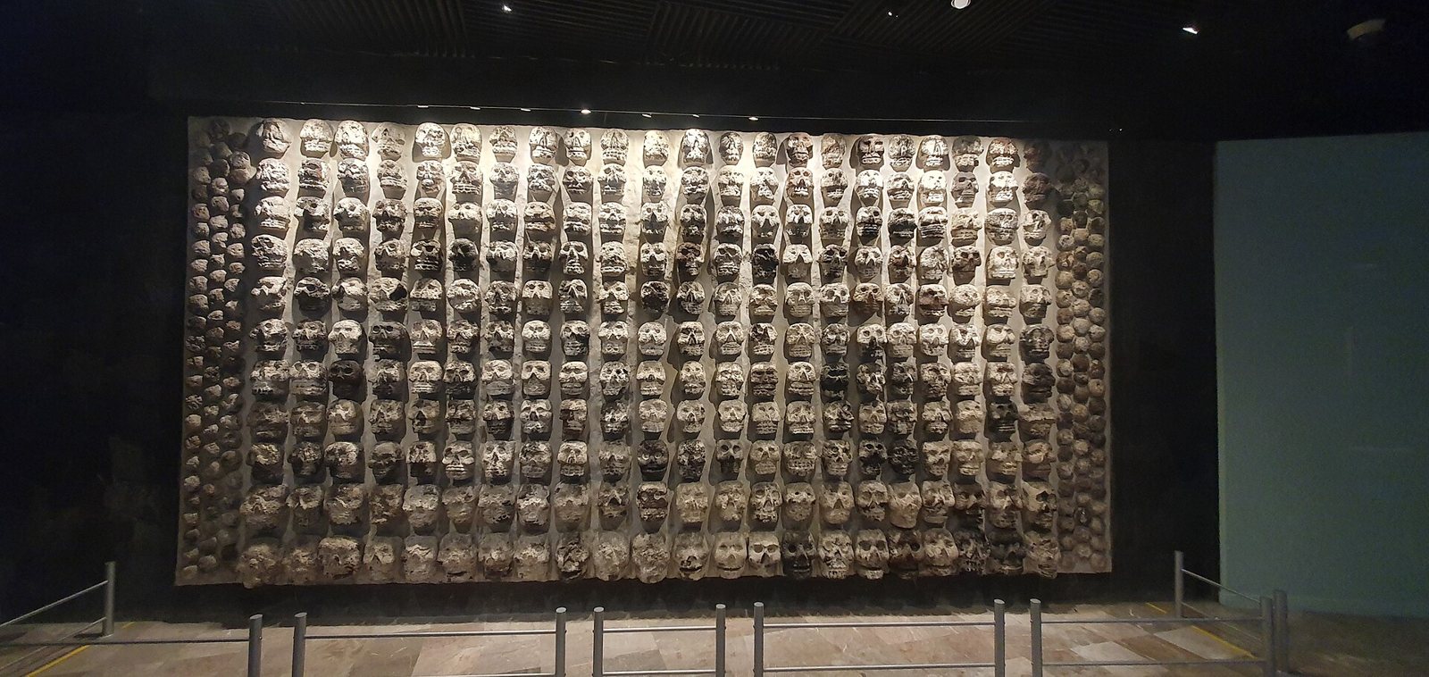 A large replica of a tzompantli wall in a dim museum gallery, hundreds of skulls set in rows behind a low barrier