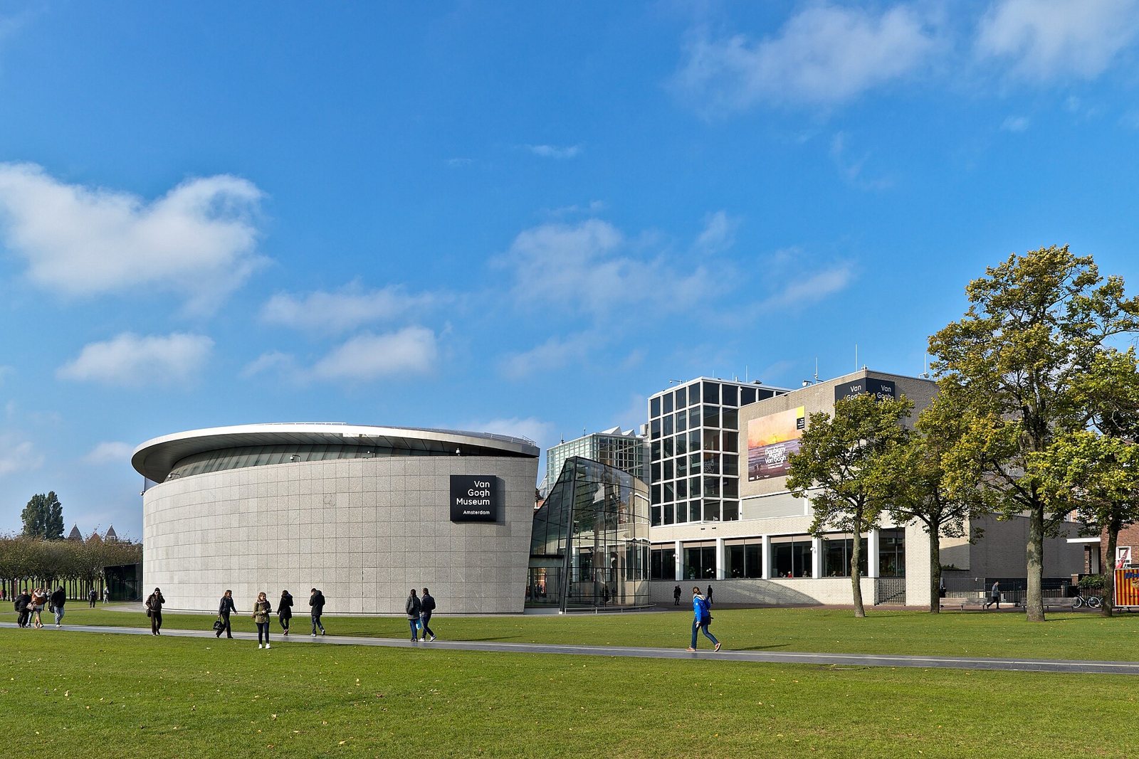 The round and angular buildings of the Van Gogh Museum seen across a lawn, with people walking on a path and trees at right