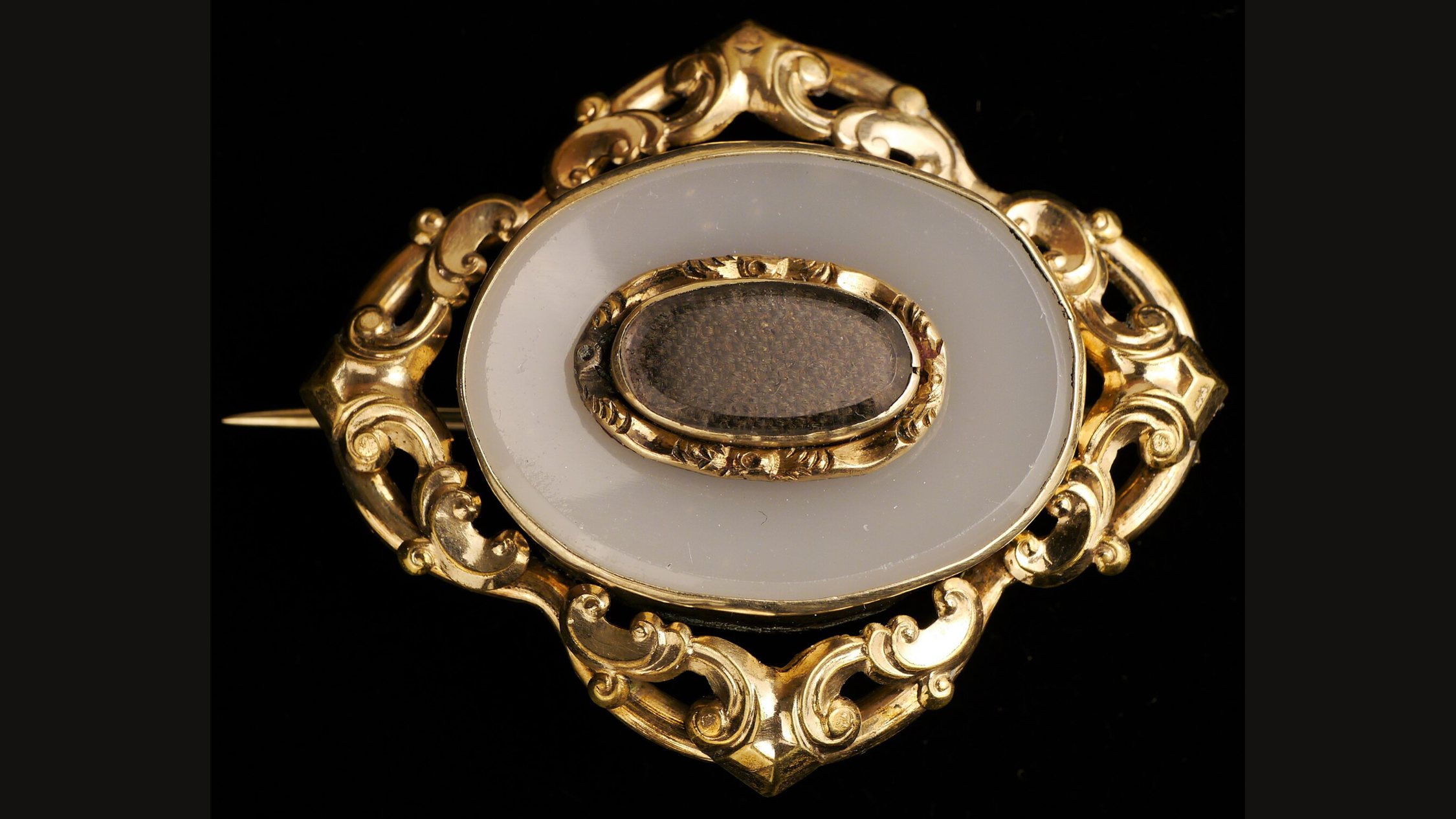 A Victorian gold brooch with a pale oval panel and a small gold frame holding a lock of hair, on a black ground
