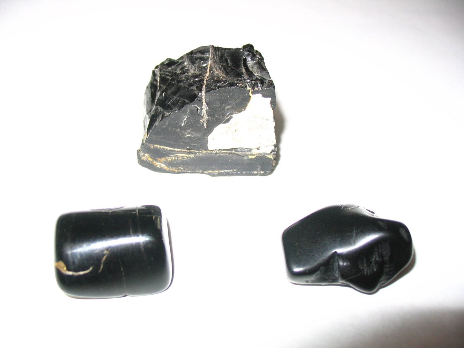 A rough black piece of jet with white mineral veins above two smooth polished black pieces, on white