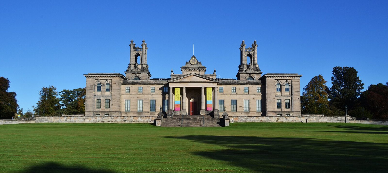 The stone front of Modern Two in Edinburgh, a classical building with a columned porch and two small towers, seen across a wide green lawn under a blue sky