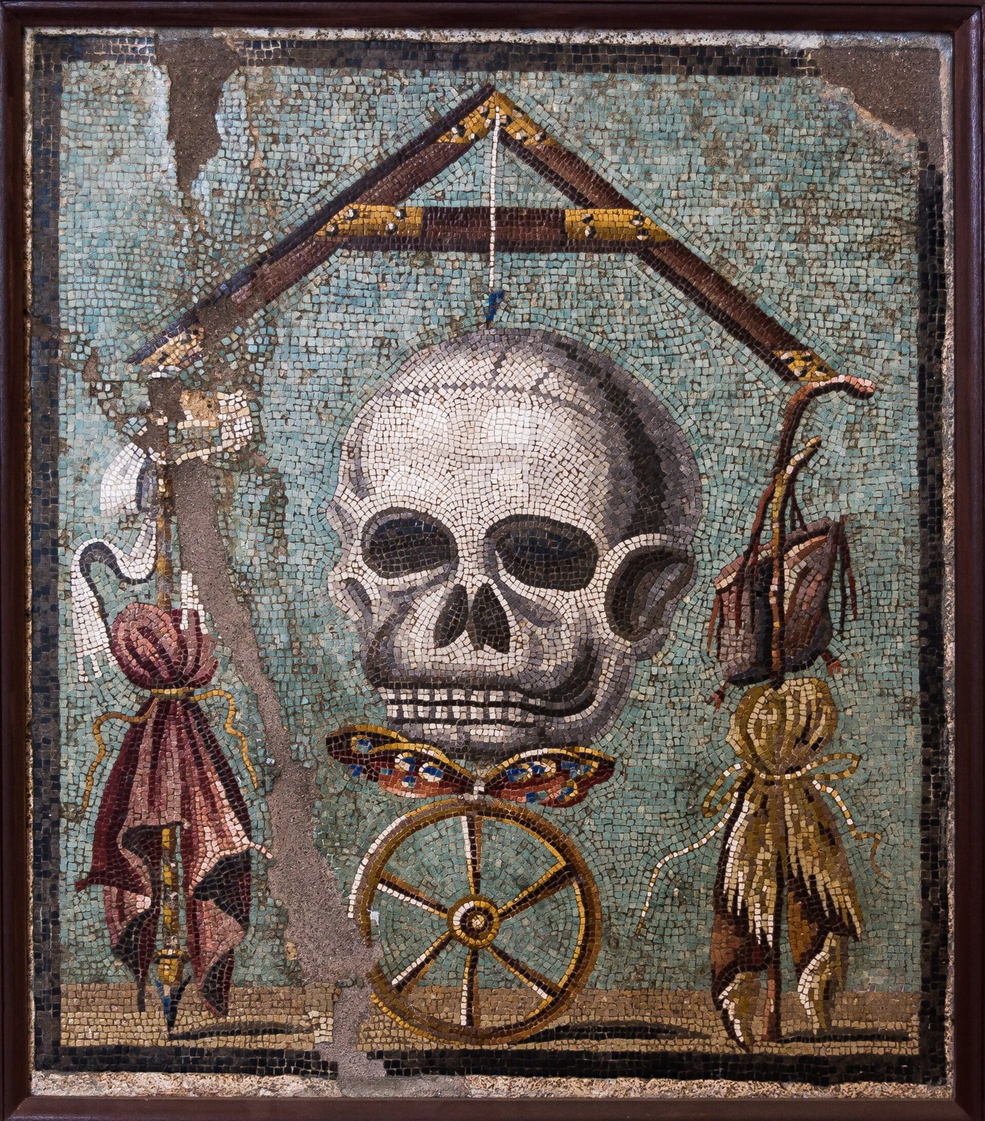 Mosaico romano de una calavera que cuelga de una plomada bajo una escuadra, sobre una mariposa y una rueda, con telas atadas a ambos lados