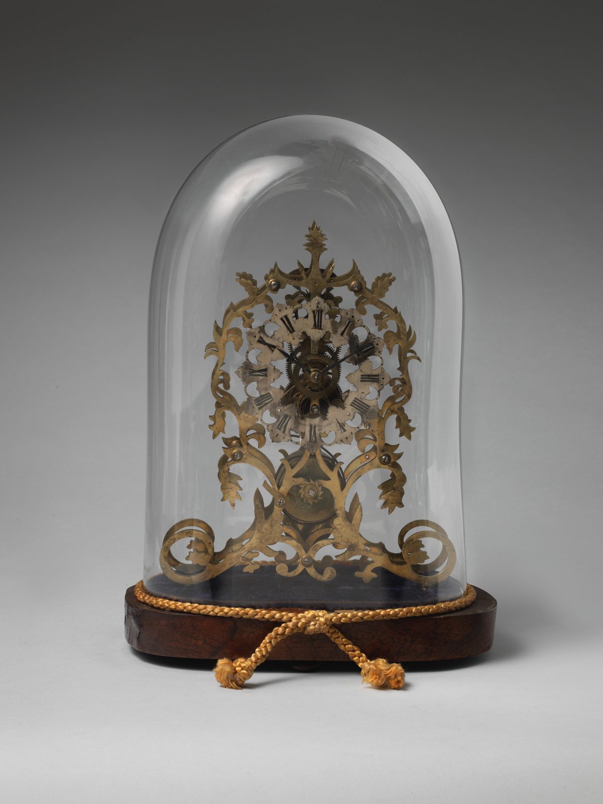 A gilded brass skeleton clock with scrolling open-work leaves around its dial, standing under a glass dome on a dark wooden base