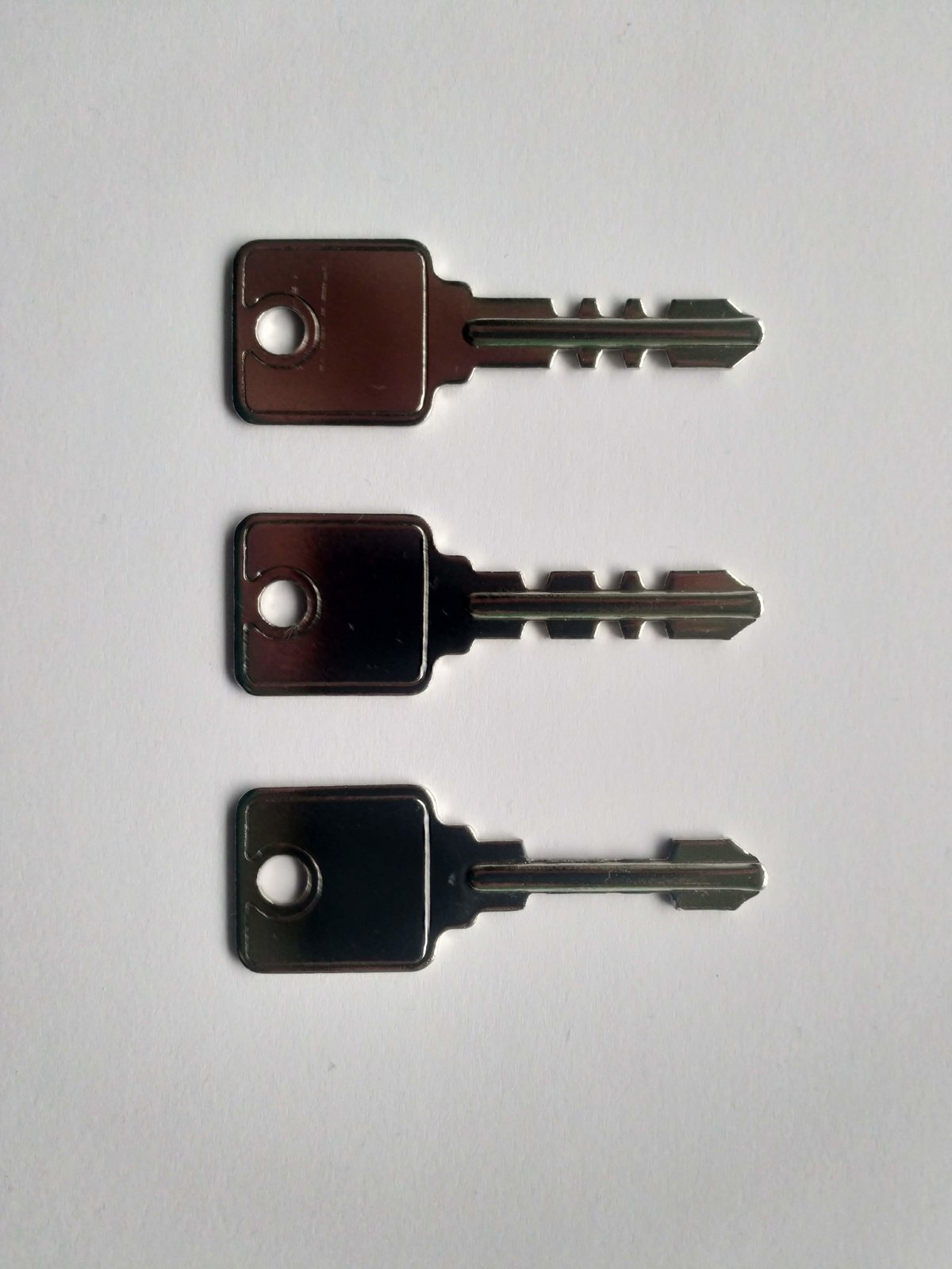 Three flat steel keys with round-hole bows laid one above another on a pale grey background: the top two have notched bits and the bottom one has a plain, smooth bit