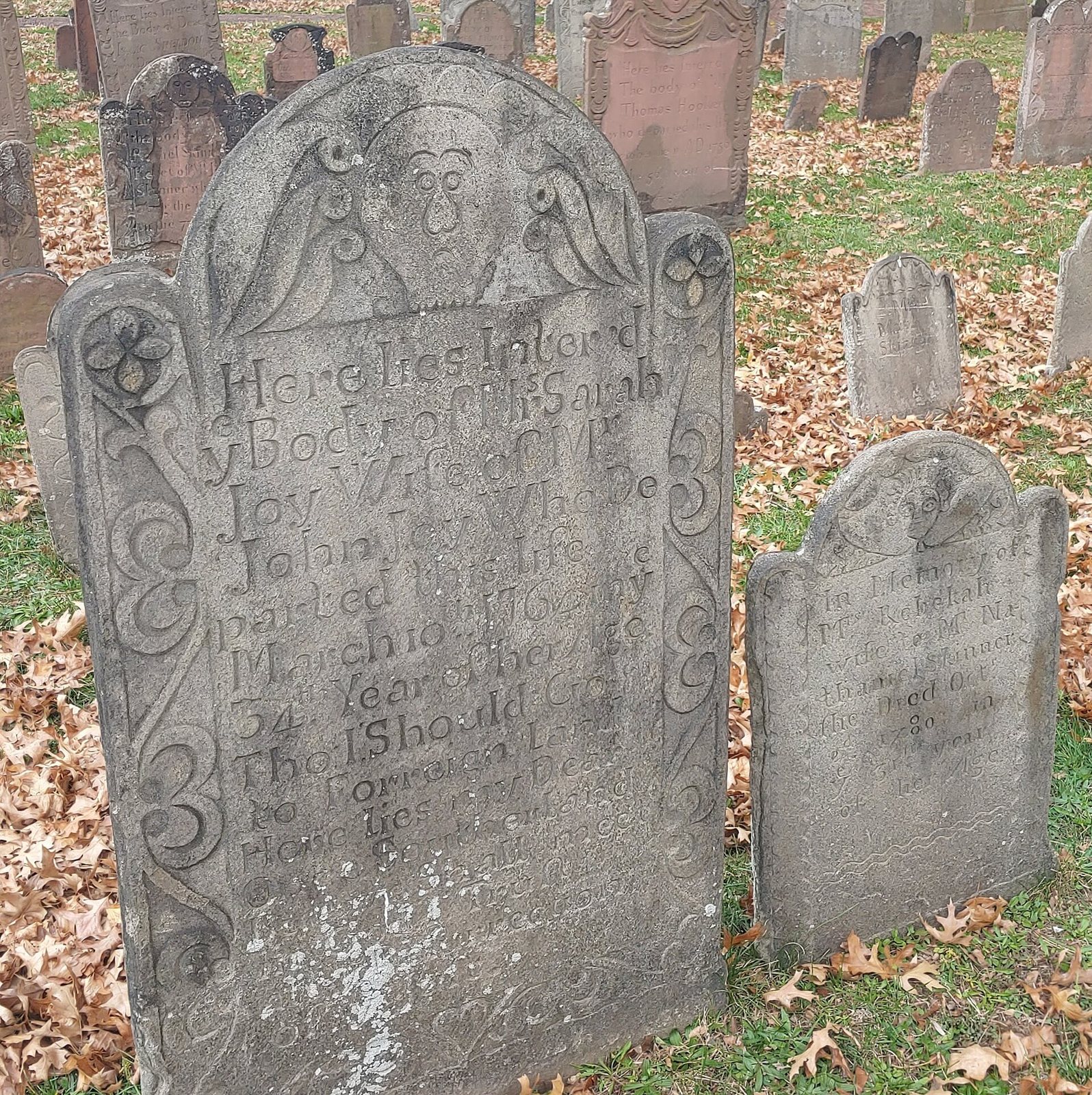 A tall brownstone gravestone with a carved winged face and scrolled side panels, a smaller stone beside it, among fallen leaves in Hartford's Ancient Burying Ground