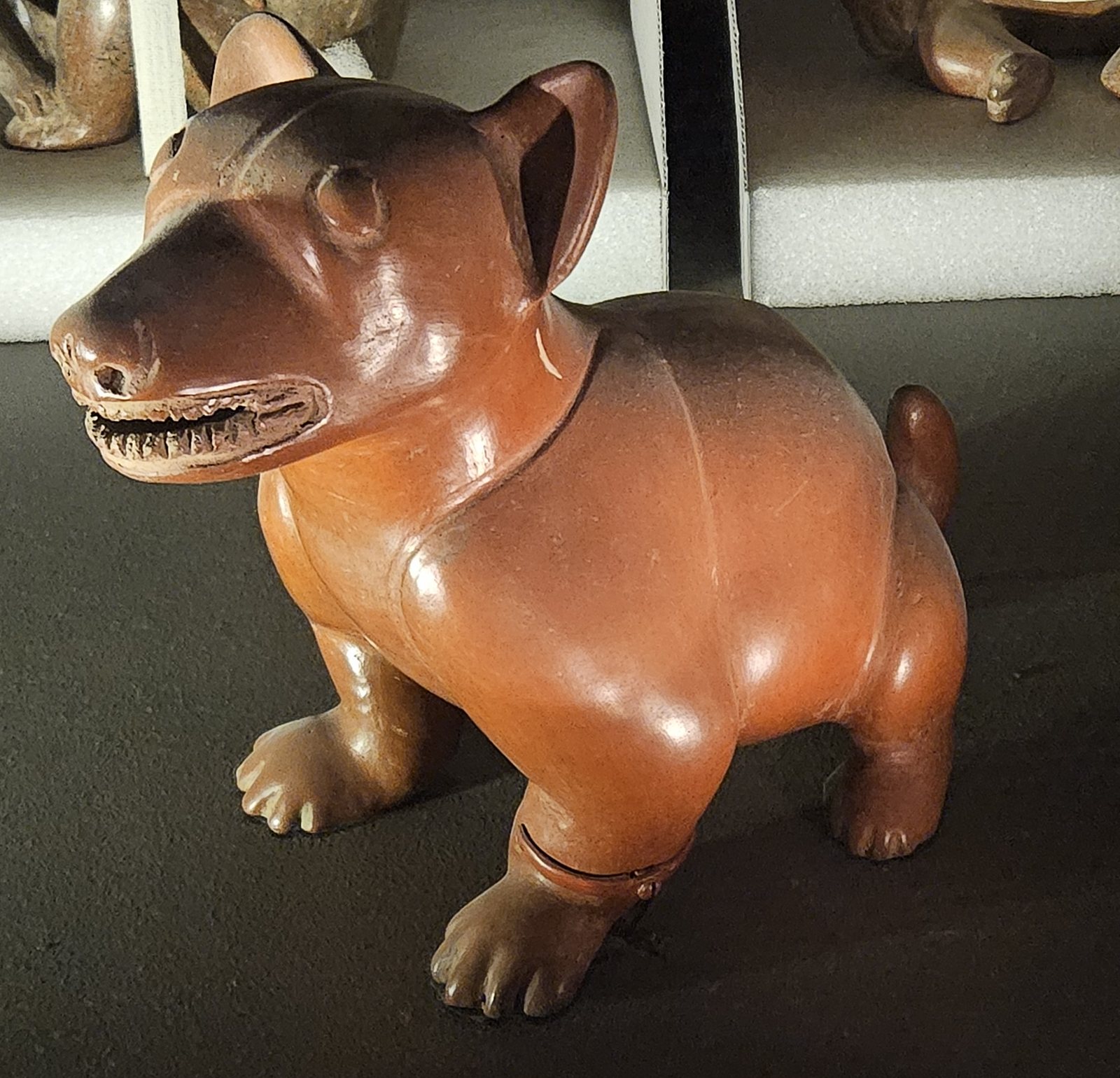 Un perro de cerámica color naranja pardo y brillante, sentado erguido, con las orejas erguidas y la boca abierta mostrando los dientes, sobre una superficie oscura