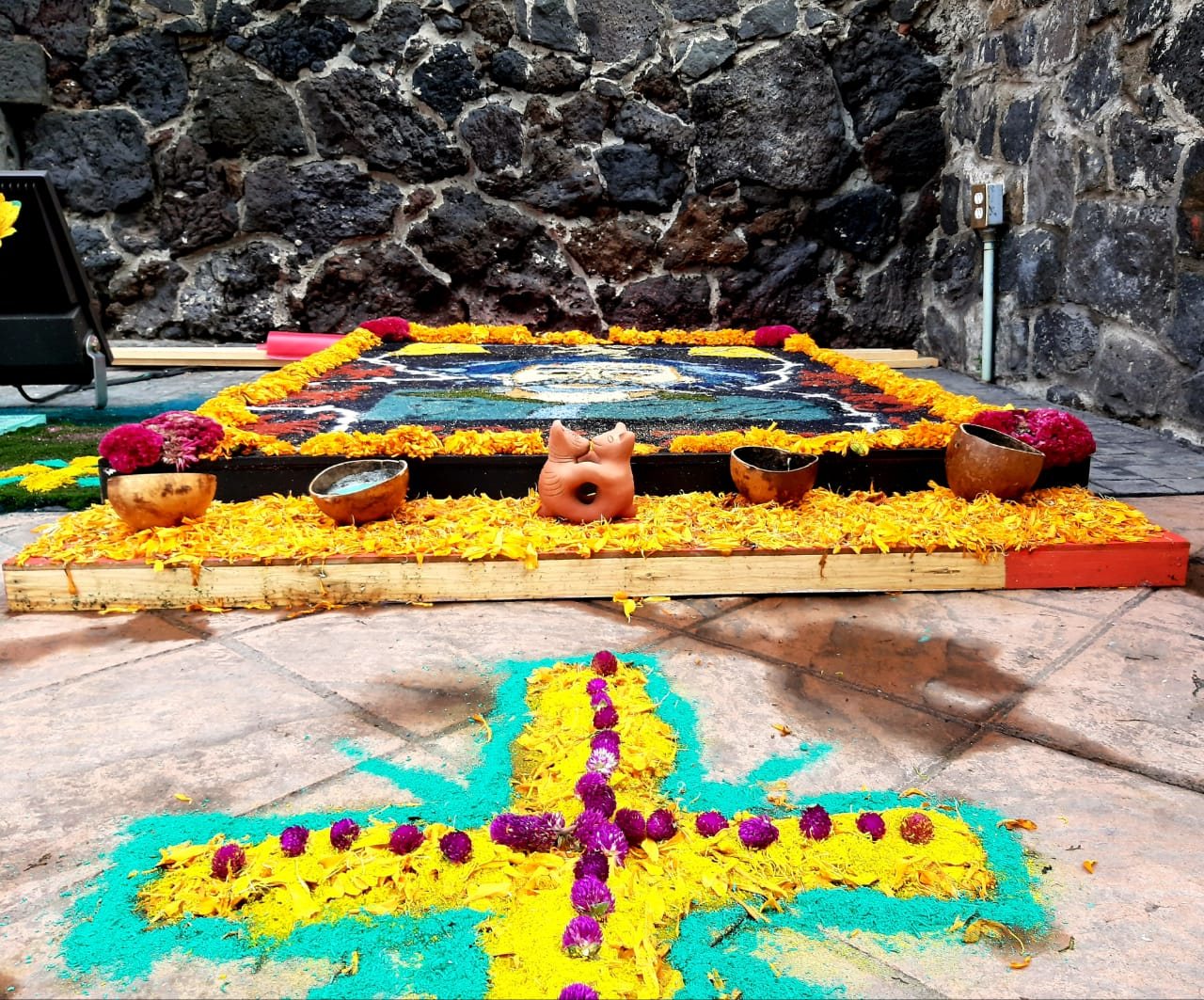 Una alfombra de Día de Muertos de aserrín de colores y pétalos de cempasúchil sobre un patio de piedra, bordeada de cempasúchil naranja y pequeños cuencos de barro, con una cruz de flores amarillas y moradas en primer plano